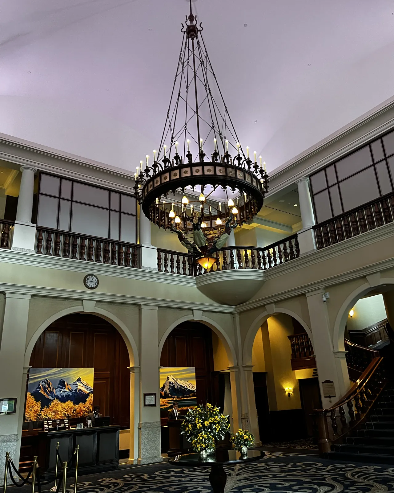 The lobby of the Fairmont Chateau Lake Louise, with a large iron candle-style chandelier hanging beneath a domed ceiling, arched doorways, mountain paintings and a curved wooden staircase