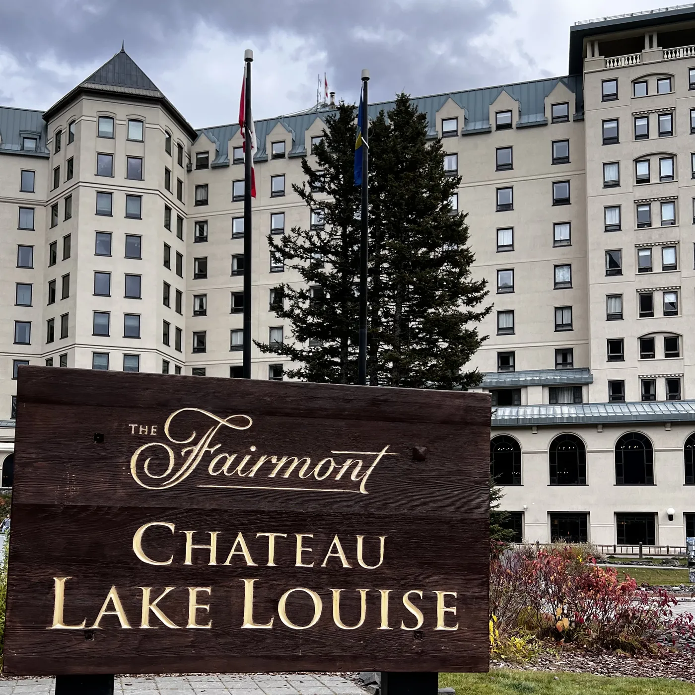 The carved wooden Fairmont Chateau Lake Louise sign in front of the hotel's cream-colored facade, with a tall evergreen and flags between the wings