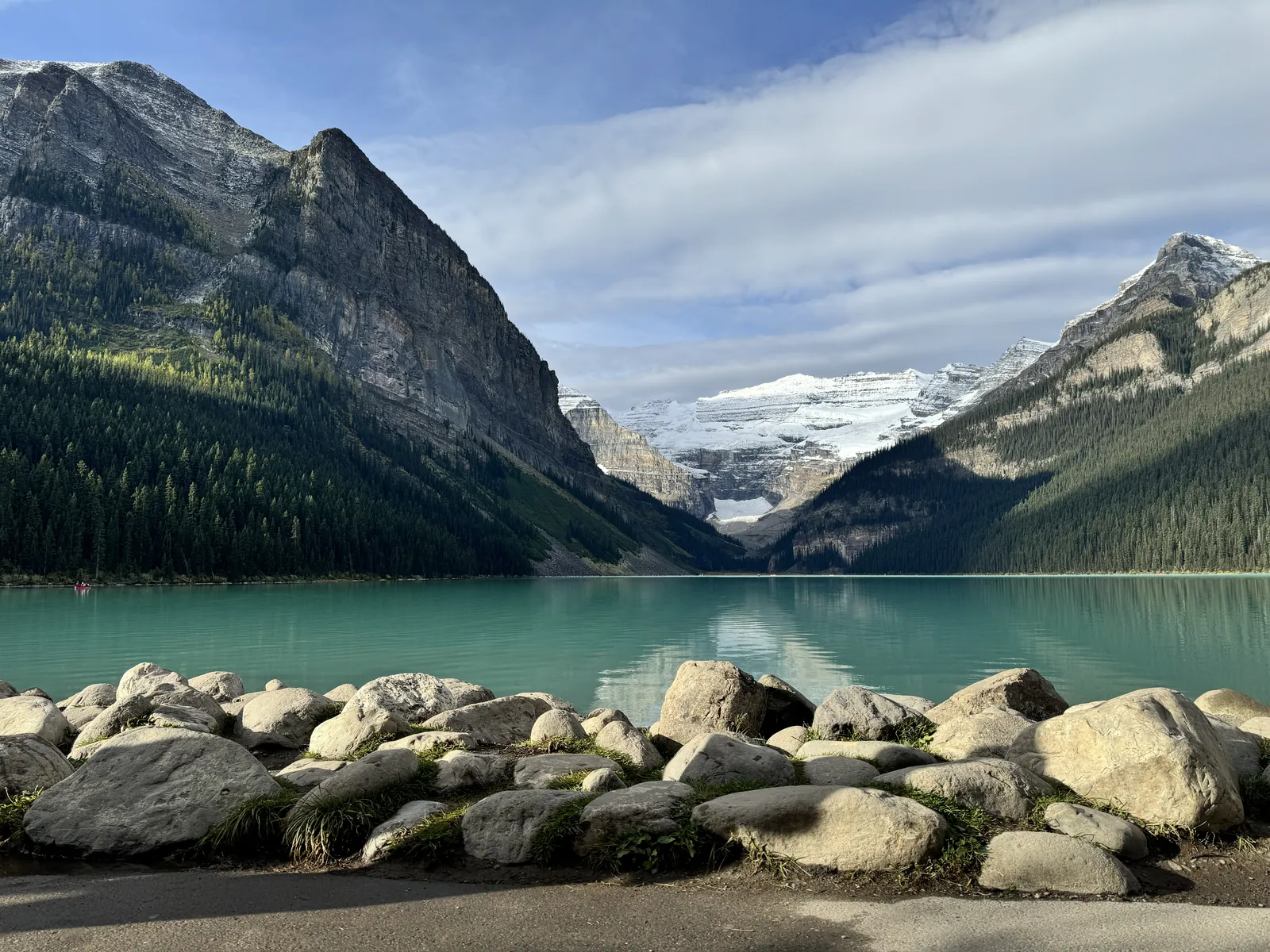 Turquoise Lake Louise below steep forested slopes with the Victoria Glacier at the far end and large boulders along the near shore