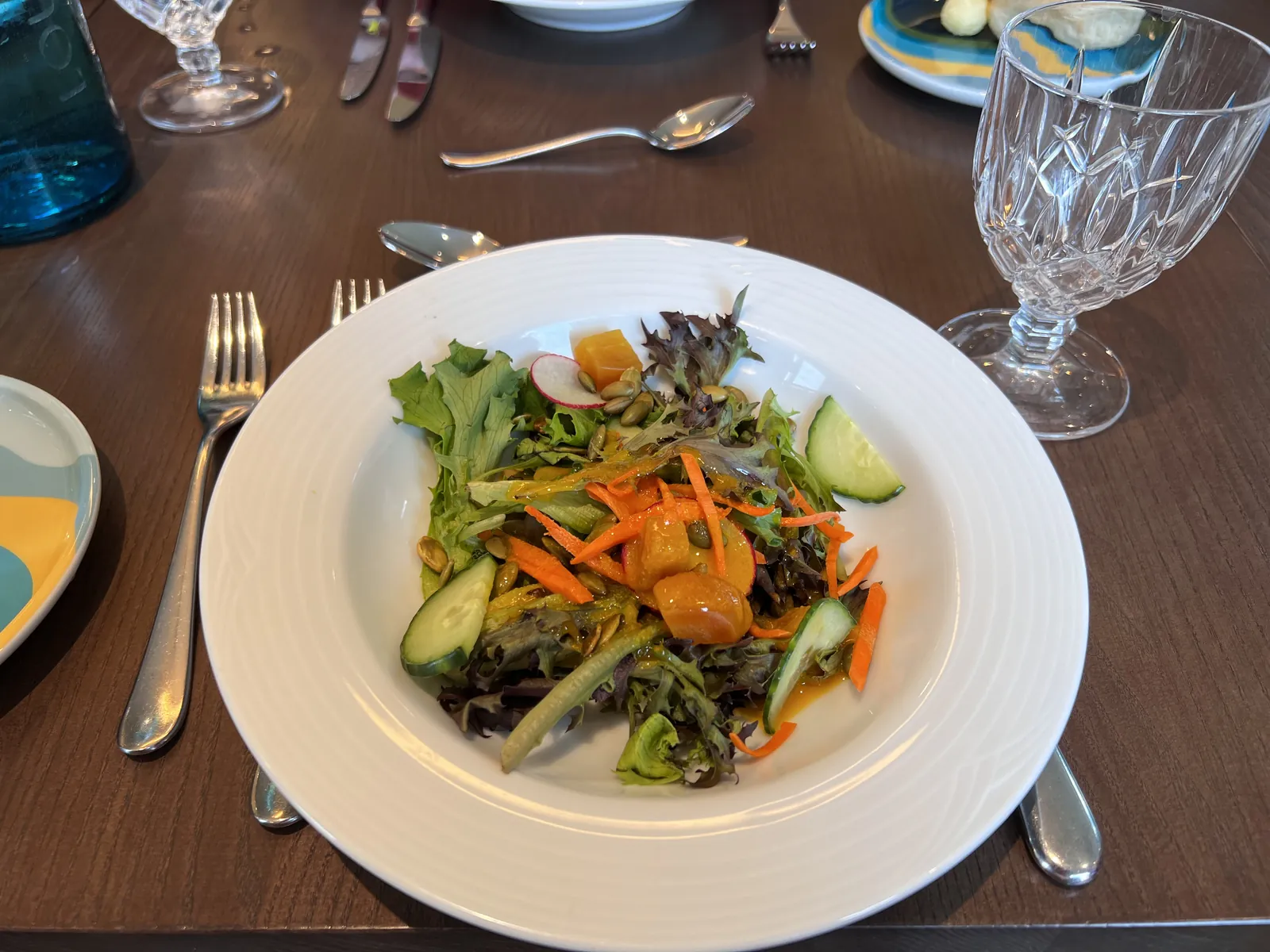 A plated green salad with carrot ribbons, cucumber, radish and squash beside crystal glassware on a dark wood table