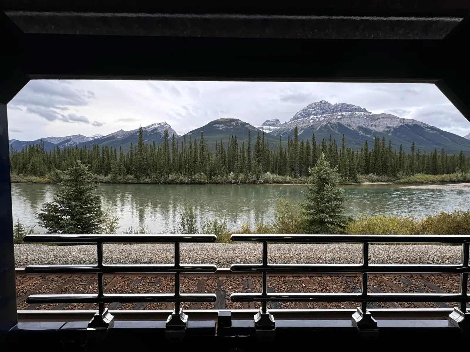 The view from the open-air vestibule at the back of a Rocky Mountaineer GoldLeaf car, rails running away past a river toward snow-dusted peaks