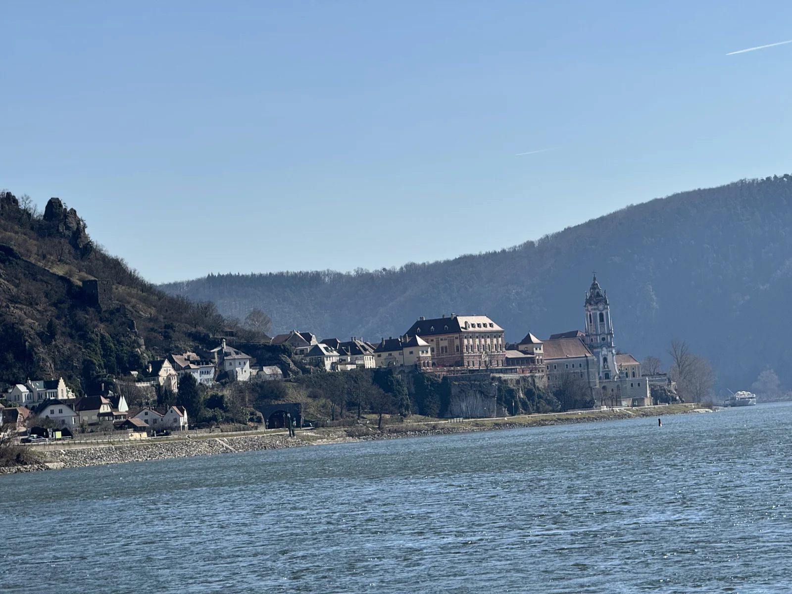 A Wachau Valley village on the Danube seen from a river cruise ship, with a church tower above the water and bare winter trees on the hillside.