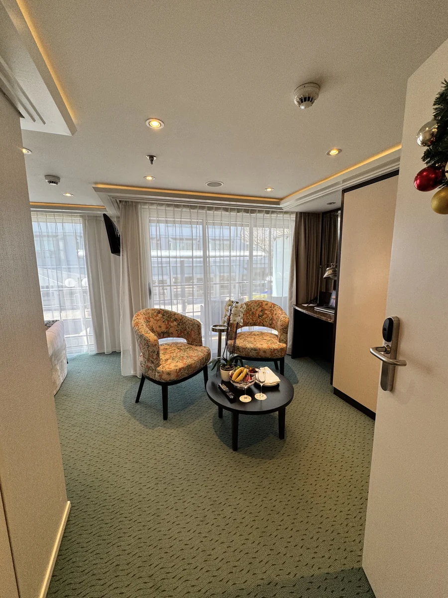 A river cruise stateroom with a small decorated Christmas tree by the door and a sliding glass door onto the river.