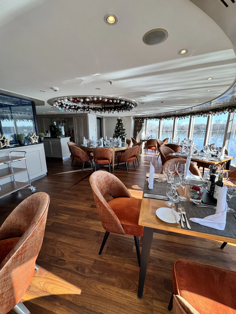 A river cruise ship dining room decorated for Christmas, with a lit tree by the windows, garlands overhead and tables set for dinner beside the river.