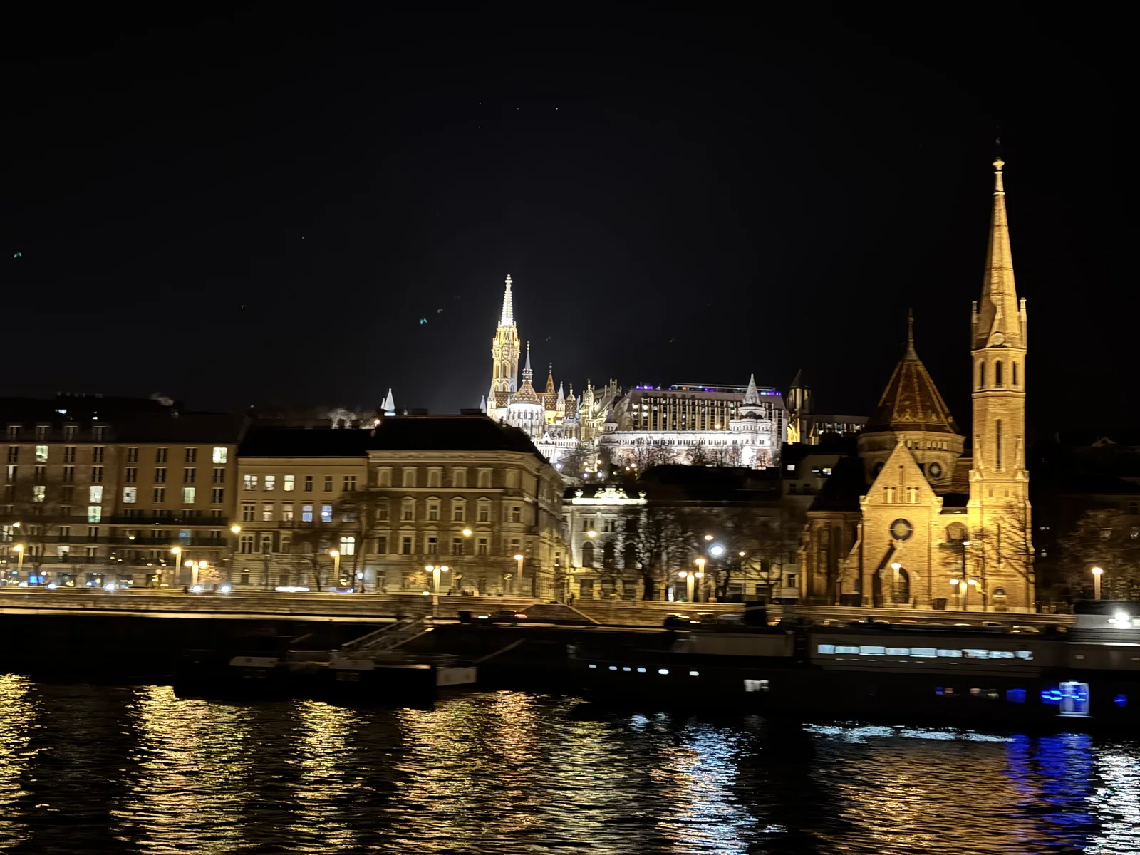 Budapest lit up at night seen from the Danube, with Matthias Church and Fisherman's Bastion glowing on the hill above the river.