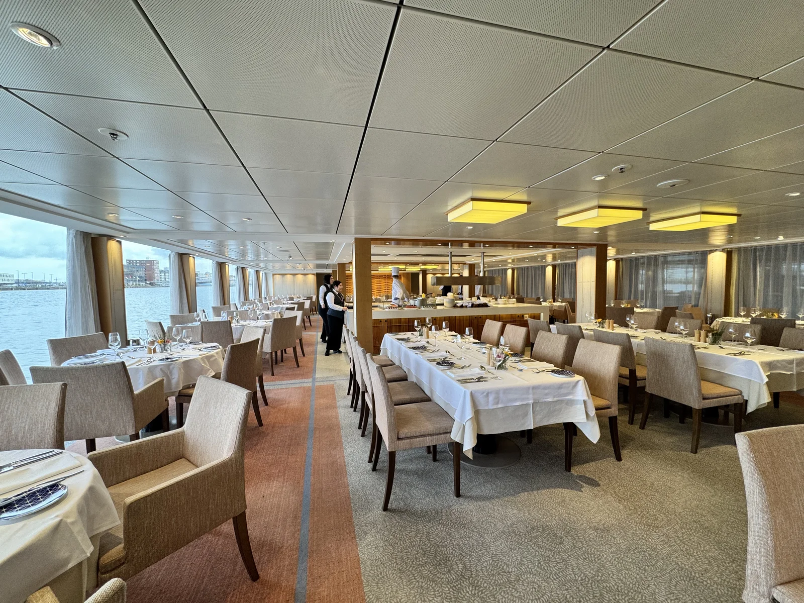 The dining room on a river cruise ship, tables set with white linens beside windows facing the river.