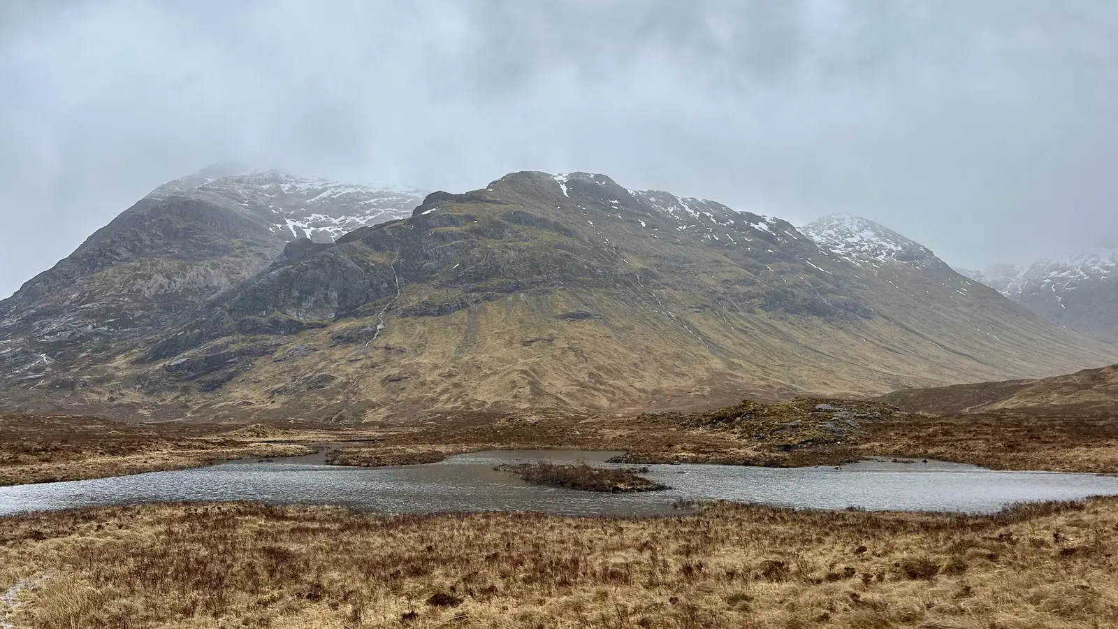 A remote glen in the Scottish Highlands, a small lochan below rugged, snow-flecked mountains near Glencoe